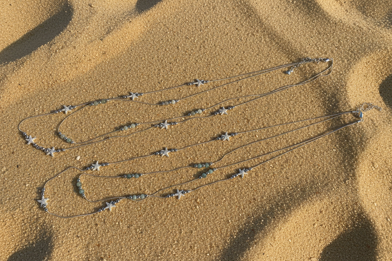 A long silver-toned beaded necklace with starfish charms and small gray beads, laid out on a sand surface.