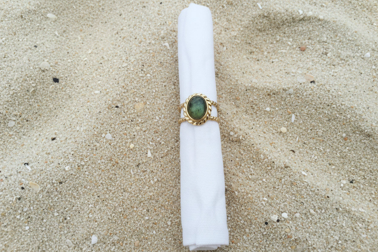 A gold ring with an oval green stone, displayed on a white fabric ring holder against a textured sand background.