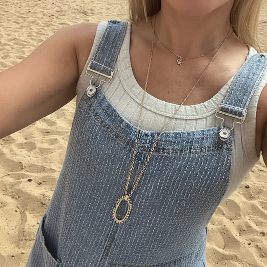 A close-up of a person wearing light blue denim overalls over a white ribbed tank top, accessorized with layered gold necklaces.