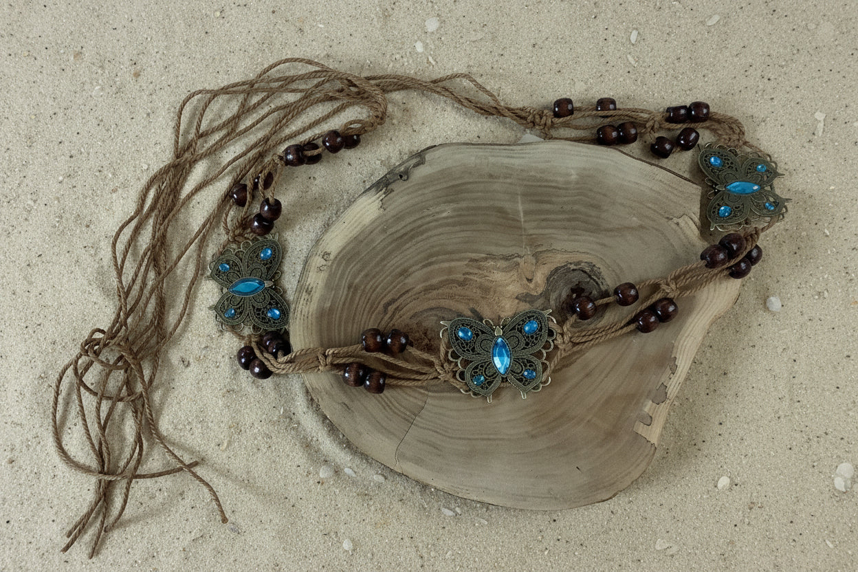 A bohemian-style macrame necklace with turquoise and dark brown beads, displayed on a rustic wood slice against a sand background.