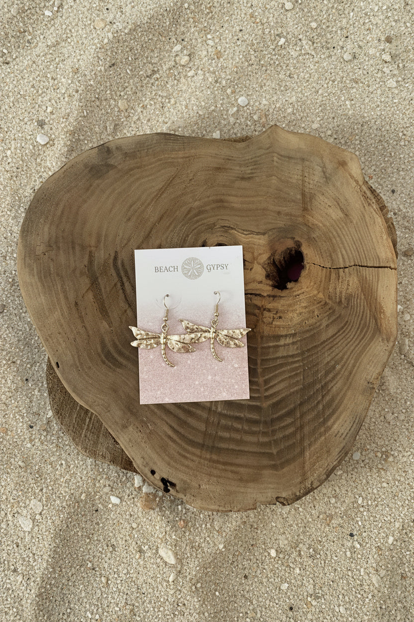 A pair of gold-toned dangle earrings with clear rhinestone details, displayed on a white card labeled 'BEACH GYPSY' and placed on a round natural wood slice against a bright pink background.