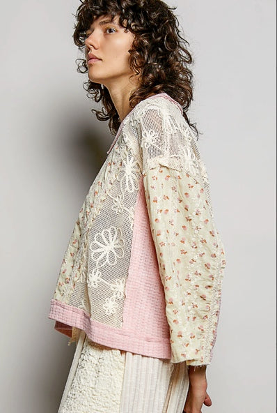 A model wearing a layered bohemian-style top featuring cream floral lace, white floral embroidery, and a pink textured inner layer, paired with cream pleated trousers, set against a plain gray studio background.