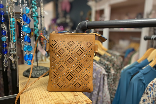 A tan perforated leather crossbody bag with silver studs, displayed on a wooden table in a retail store setting with clothing racks and jewelry in the background.