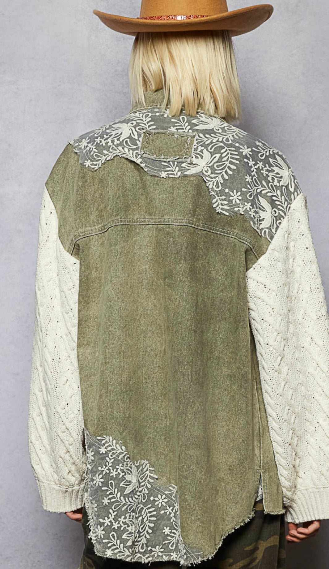 Person wearing a green denim vest with white lace details and a brown hat on a gray background