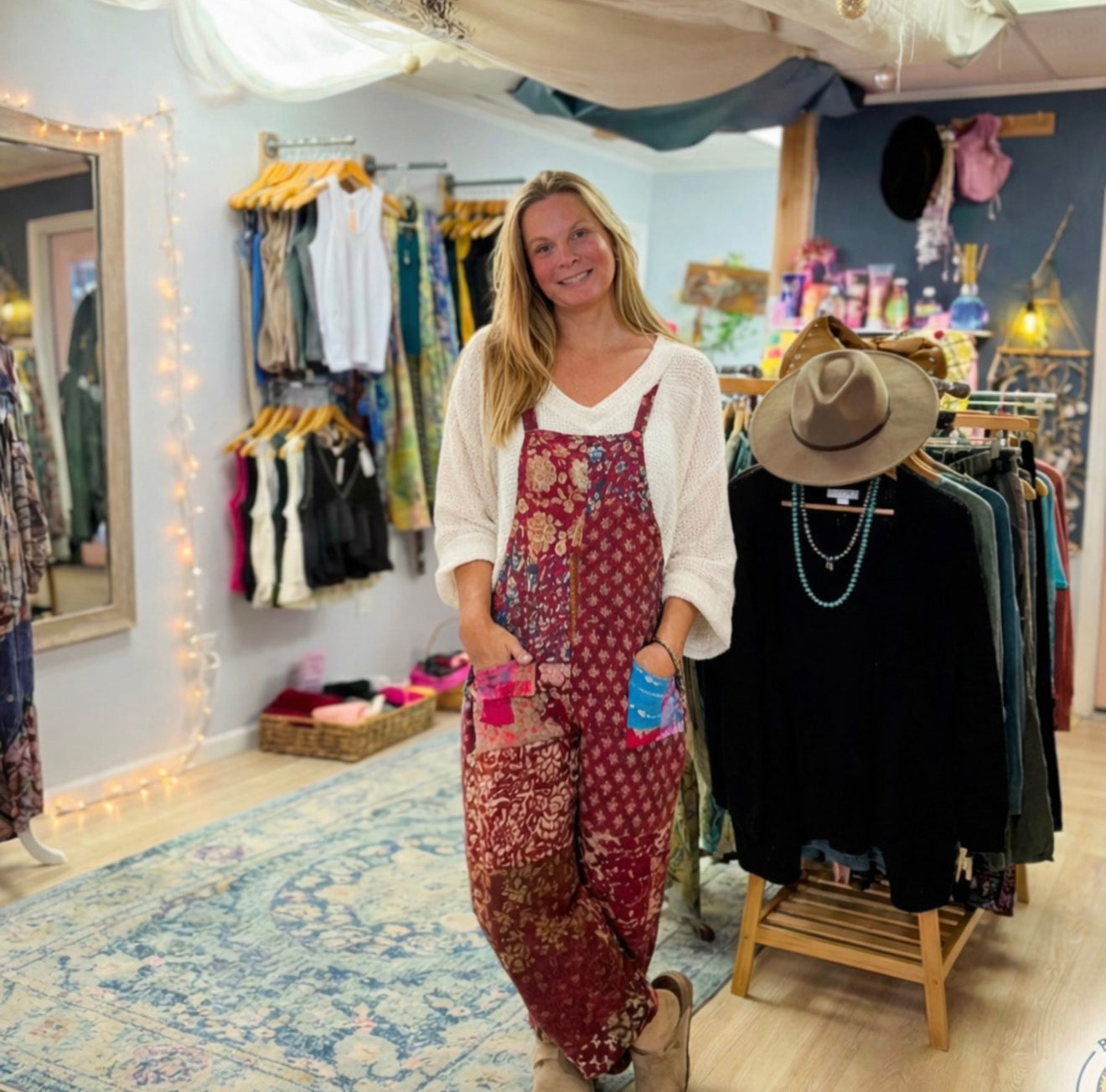 Woman in a patterned jumpsuit standing in a clothing store with racks of clothes and a mirror.