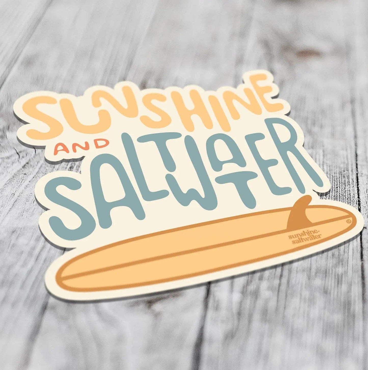 A die-cut sticker featuring the words 'SUNSHINE AND SALTWATER' in playful orange and blue lettering above a tan surfboard, shown on a light gray wood plank background.