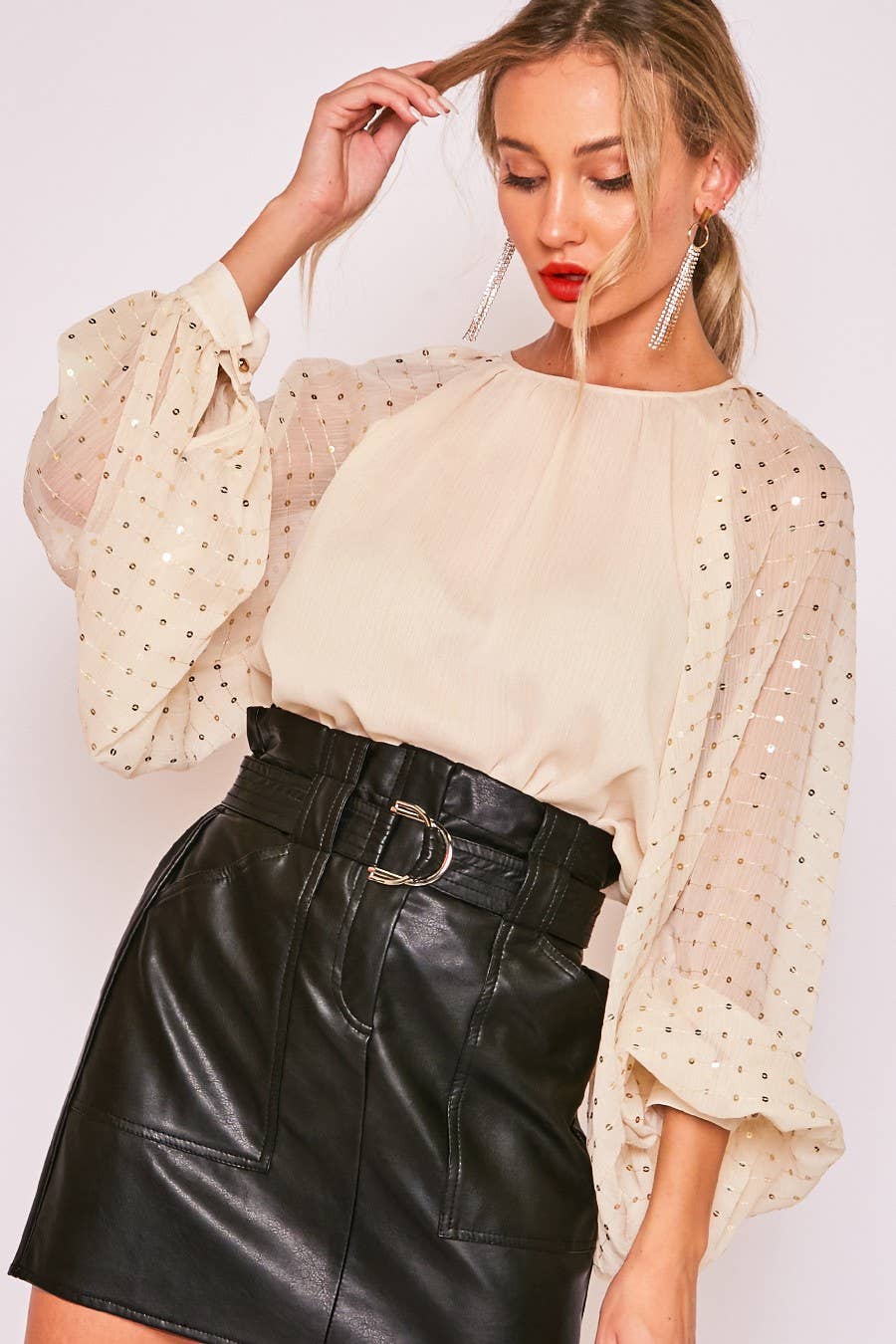 Woman wearing a beige blouse with sheer sleeves with sequins and a black leather skirt on a white background