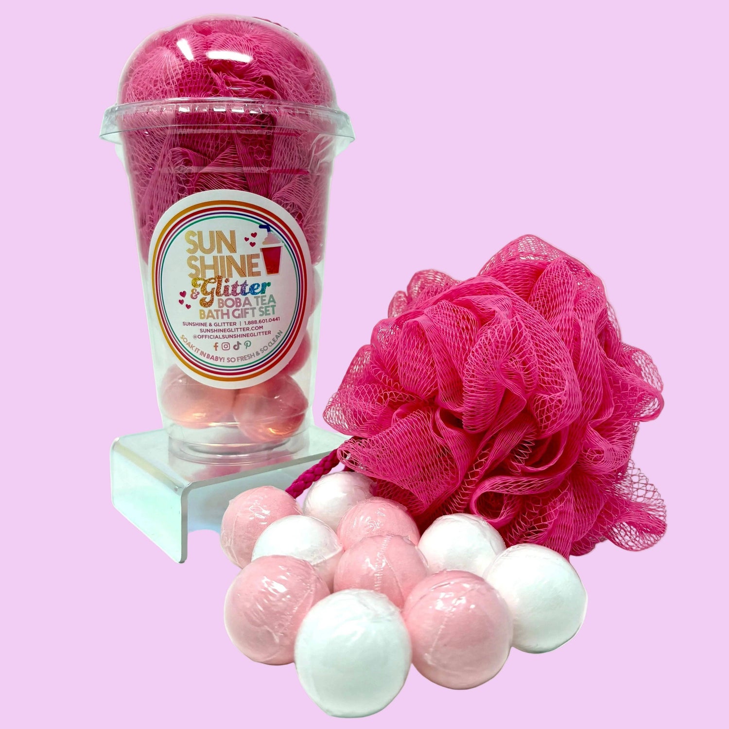 Bath gift set with pink loofah, bath bombs, and a labeled container on a pink background