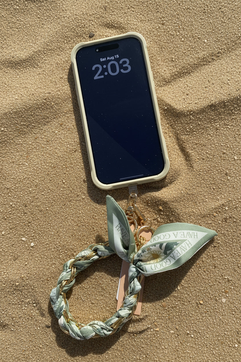 A sage green phone case with a matching silk scarf lanyard and gold-tone hardware, shown on a pink fabric background with a smartphone inserted.