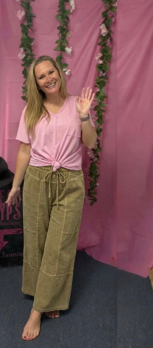 Woman in pink top and green pants waving at a pink backdrop with floral decorations.
