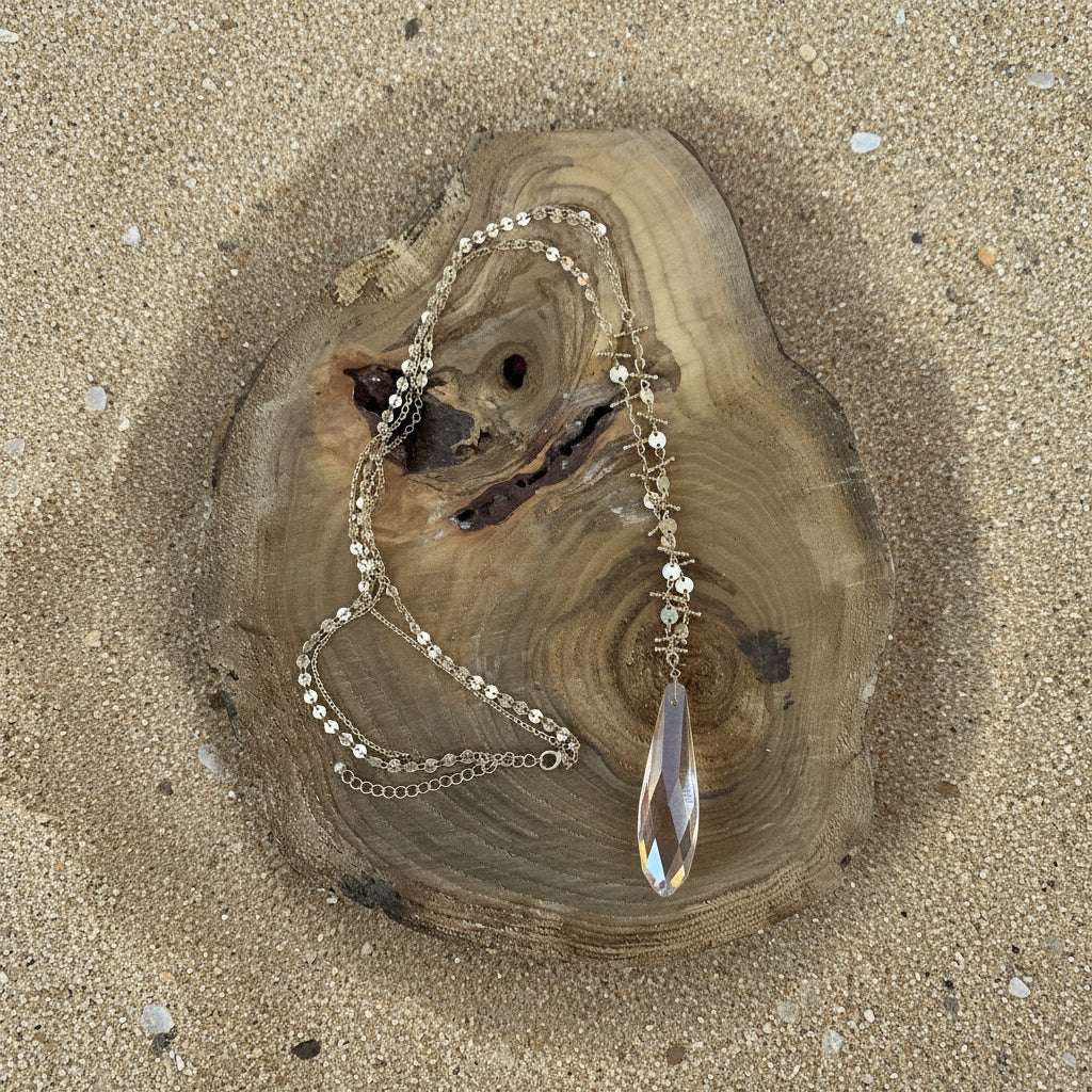A gold-toned beaded necklace with a large iridescent teardrop crystal pendant, displayed on a rustic wood slice against a bright  sand background.