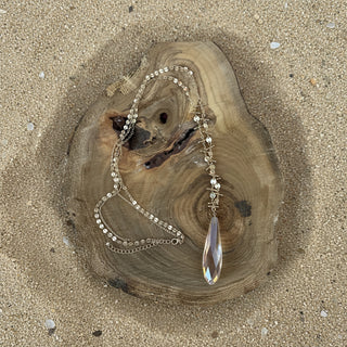 A gold-toned beaded necklace with a large iridescent teardrop crystal pendant, displayed on a rustic wood slice against a bright  sand background.