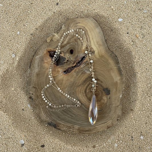 A gold-toned beaded necklace with a large iridescent teardrop crystal pendant, displayed on a rustic wood slice against a bright  sand background.