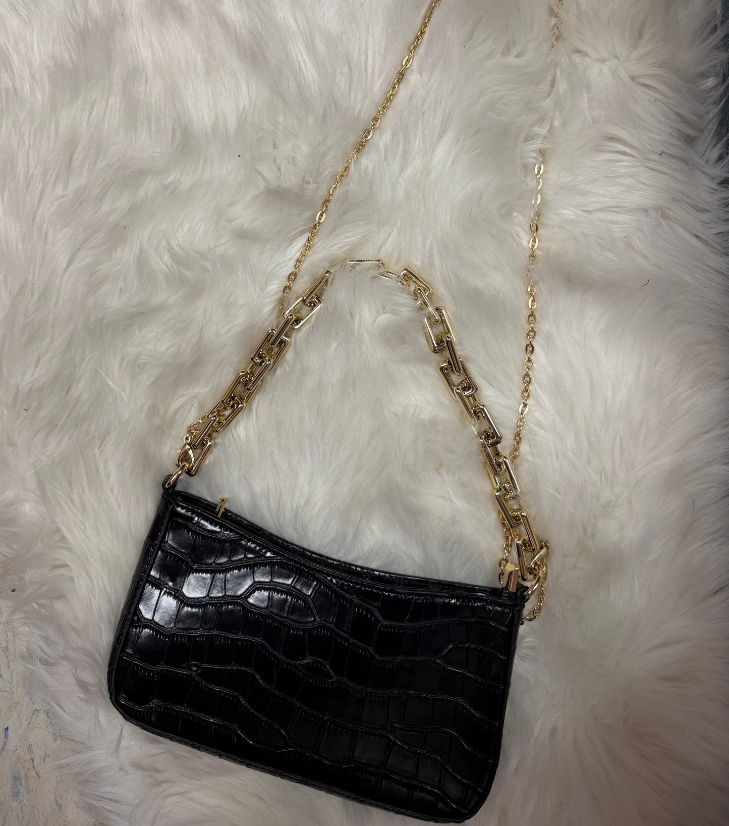 A black crocodile-embossed clutch bag with a gold chain strap, laid flat on a white faux fur surface.
