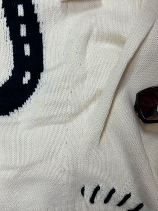 Close-up of a cream knit sweater with a dark navy road-sign pattern and black stitched detail, with a pair of sunglasses resting on the sleeve.
