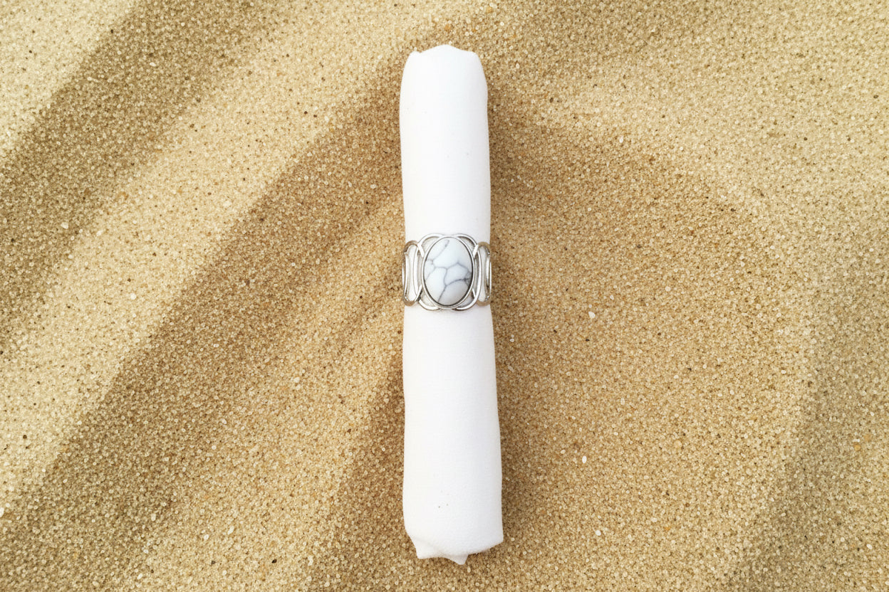 A silver ring with a large oval cabochon stone featuring a black and white marbled pattern, displayed on a white fabric ring holder against a sand background .
