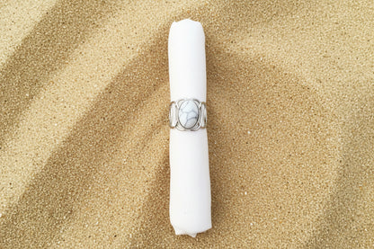 A silver ring with a large oval cabochon stone featuring a black and white marbled pattern, displayed on a white fabric ring holder against a sand background .