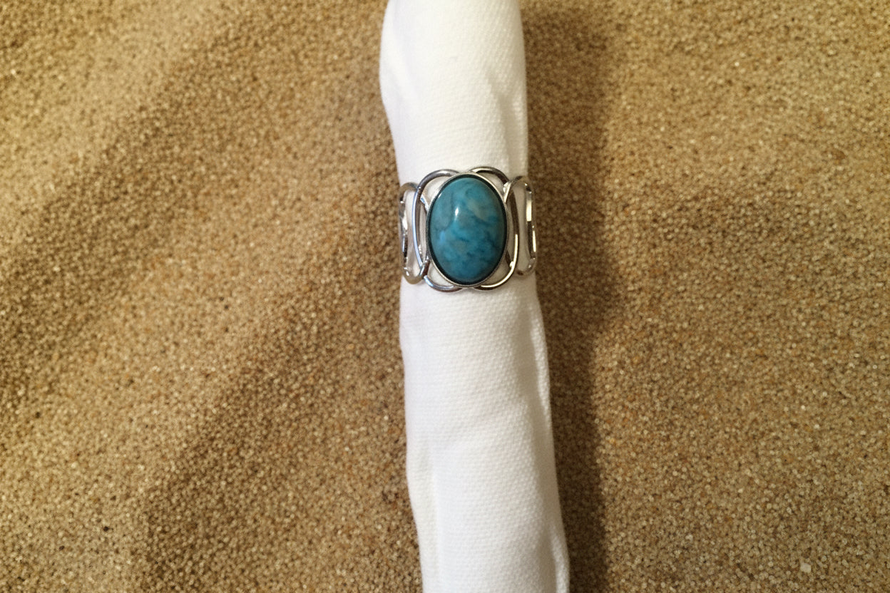 A silver ring with an oval turquoise-blue stone, displayed on a white paper tube against a sand background.
