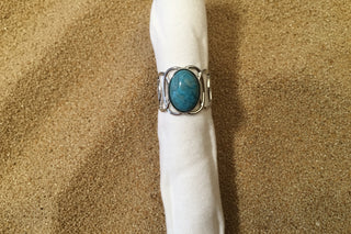 A silver ring with an oval turquoise-blue stone, displayed on a white paper tube against a sand background.