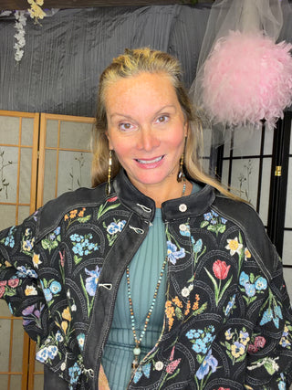 A woman wearing a black embroidered bomber jacket with colorful floral and butterfly patterns over a sage green ribbed top, standing indoors in front of a gray curtain and wooden screen.