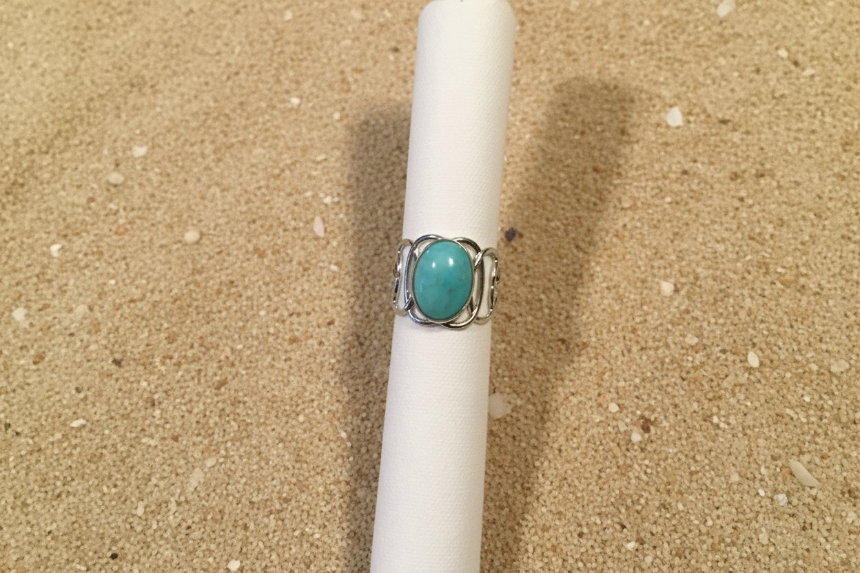 A silver ring with an oval turquoise-colored stone, displayed on a white fabric ring holder against a brown woven textile background.