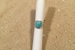 A silver ring with an oval turquoise-colored stone, displayed on a white fabric ring holder against a brown woven textile background.
