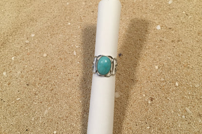 A silver ring with an oval turquoise-colored stone, displayed on a white fabric ring holder against a brown woven textile background.