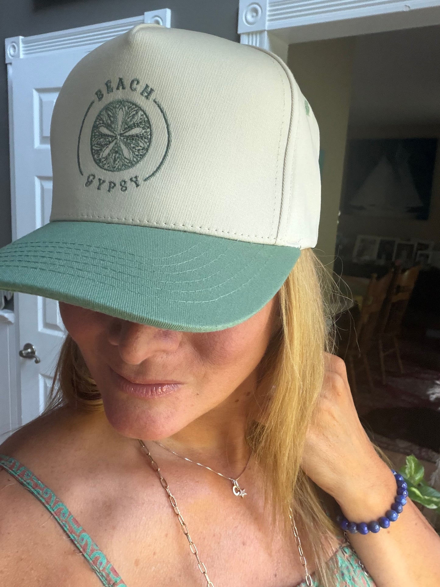 A beautiful woman wearing a cream and sage green snapback trucker hat with a circular 'Beach Gypsy' logo on the front, photographed in an indoor setting.
