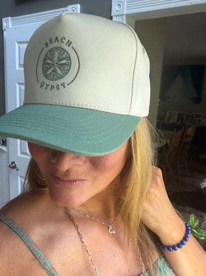 A beautiful woman wearing a cream and sage green snapback trucker hat with a circular 'Beach Gypsy' logo on the front, photographed in an indoor setting.