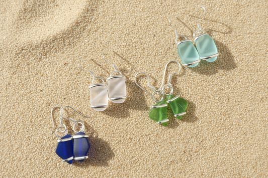 Sets of blue, green, light blue and pink and silver sea glass earrings on a sandy textured surface