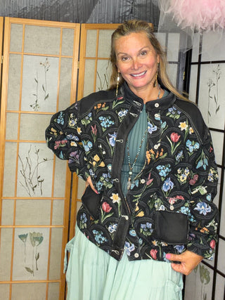 A woman posing in a black bomber jacket with a colorful floral print, a teal top, and a light green skirt, standing in front of a decorative folding screen.
