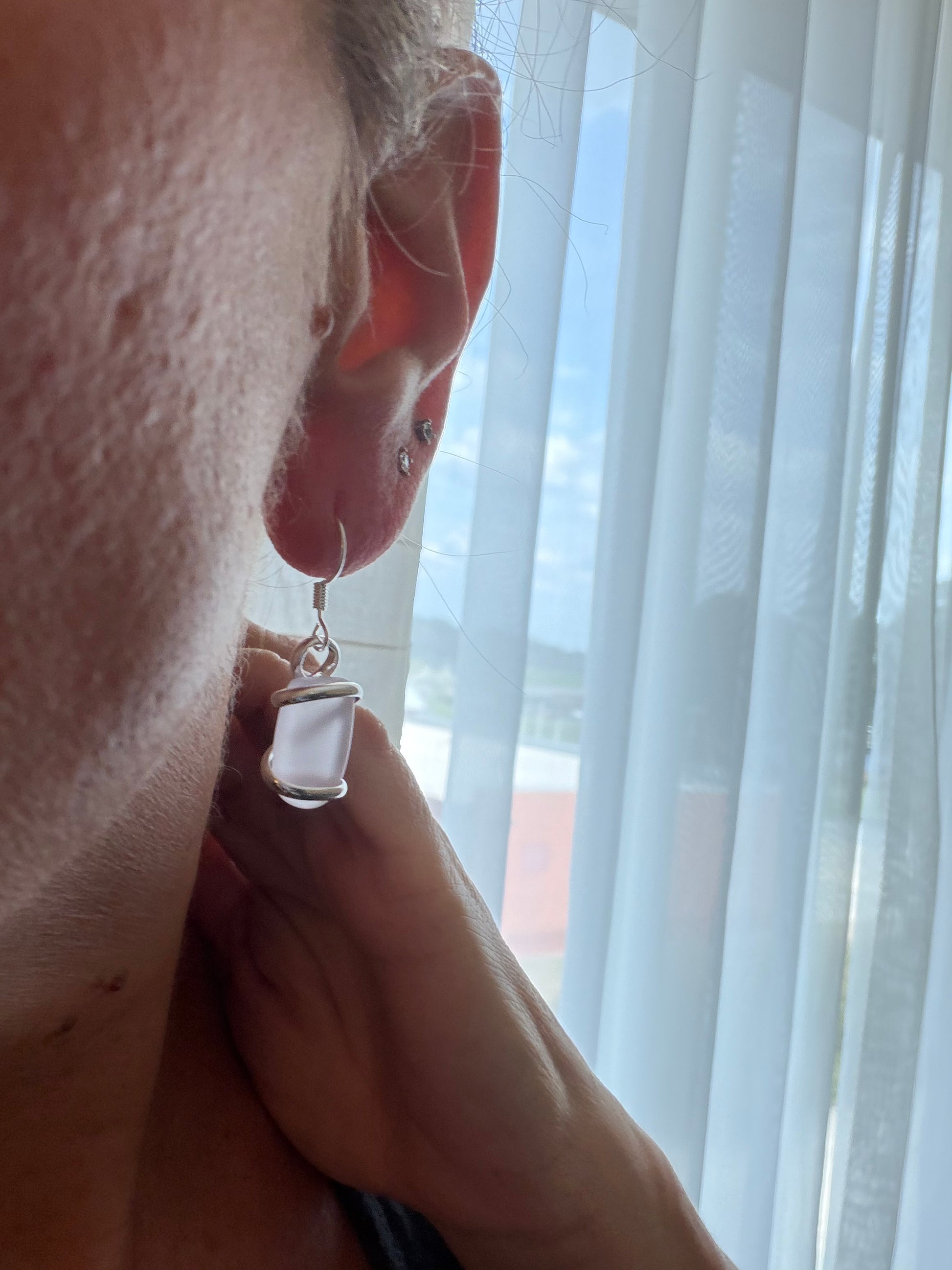 Close-up of a person wearing a silver earring with a pink sea glass, against a light curtain background 