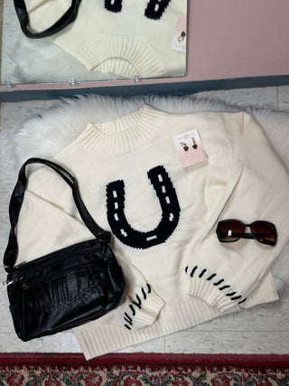 An off-white knit sweater with a large black horseshoe graphic on the chest, styled in a flat lay with a black shoulder bag, sunglasses, and a pair of earrings on a white faux fur surface.
