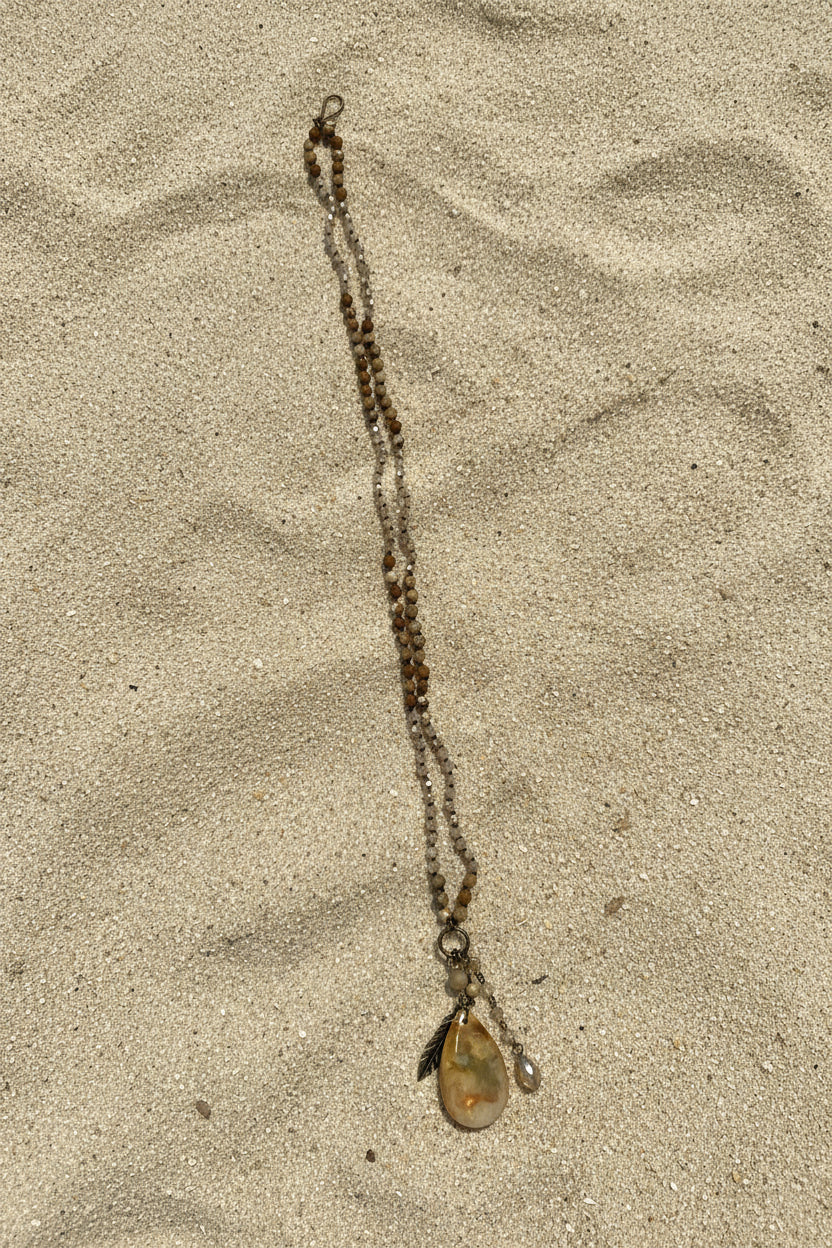 A beaded necklace with a polished oval pendant with a sand background.