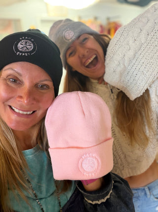 Two women smiling in a shop setting, wearing knit beanies.