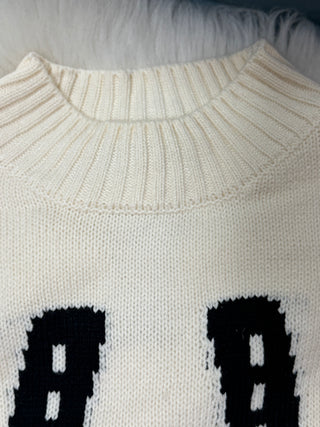 Close-up of a cream-colored knit sweater with a ribbed crew neck and large black knit lettering or numbers at the bottom.