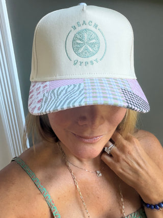 A woman wearing a Beach Gypsy brand trucker hat with a cream crown, a pastel multicolor patterned visor, and a mint green embroidered logo on the front.
