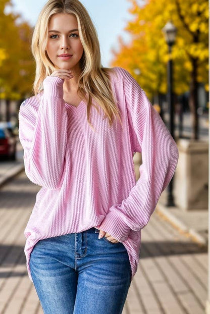 Woman wearing a pink sweater and blue jeans standing outdoors with trees in the background.