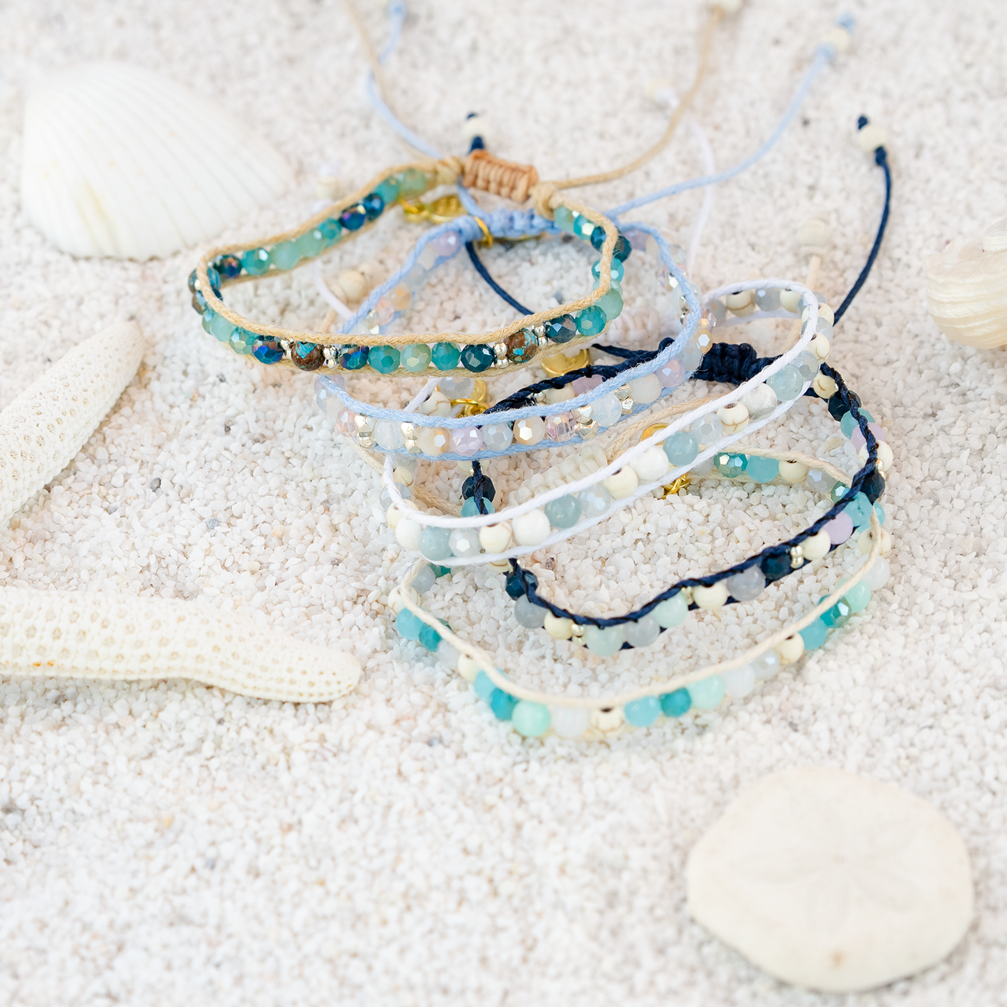 Stack of beaded bracelets on a sandy surface with seashells.