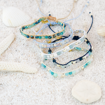 Stack of beaded bracelets on a sandy surface with seashells.