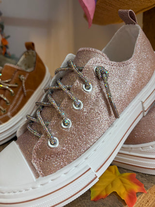 A close-up of a glittery rose gold low-top sneaker with iridescent laces and silver metal eyelets, shown alongside another tan sneaker in a styled setting with a yellow leaf prop.