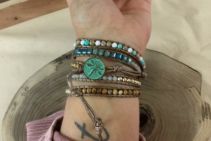 A close-up of a person's wrist wearing a stack of bohemian-style multi-strand bracelets, including a green palm tree medallion, on a rustic wood slice with a sandy background.