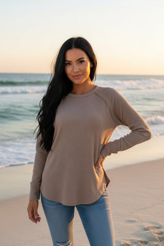 A woman wearing a long-sleeve crewneck top in a warm taupe or cocoa brown color, paired with blue jeans, against a beach background.