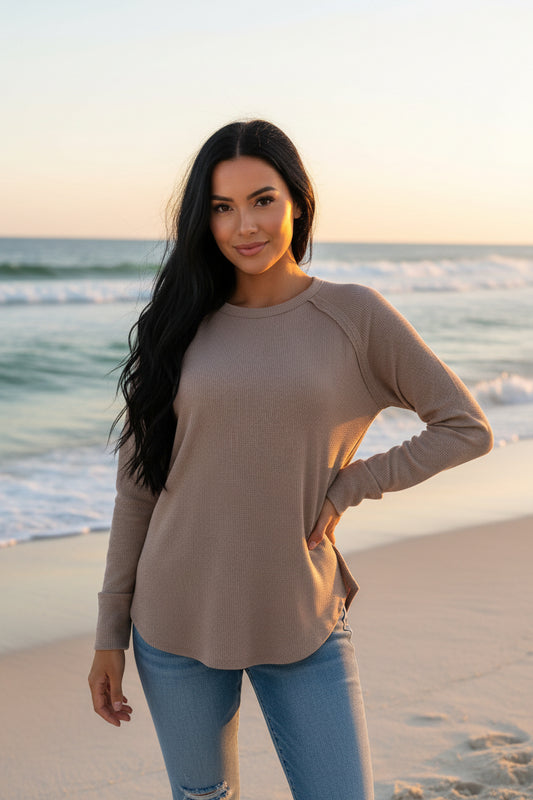 A woman wearing a long-sleeve crewneck top in a warm taupe or cocoa brown color, paired with blue jeans, against a beach background.
