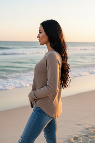 A woman in a beige long-sleeve top and blue jeans standing on a sandy beach at sunset, shown in profile.