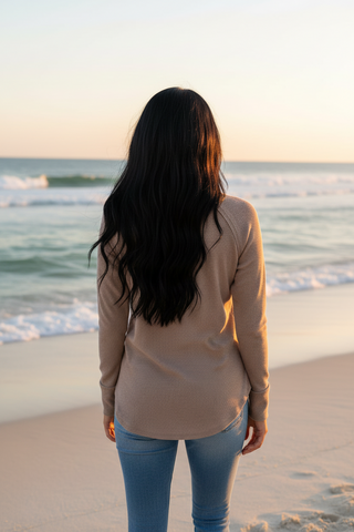 A woman with long dark hair stands on a sandy beach facing the ocean, wearing a beige long-sleeve top and light blue jeans.