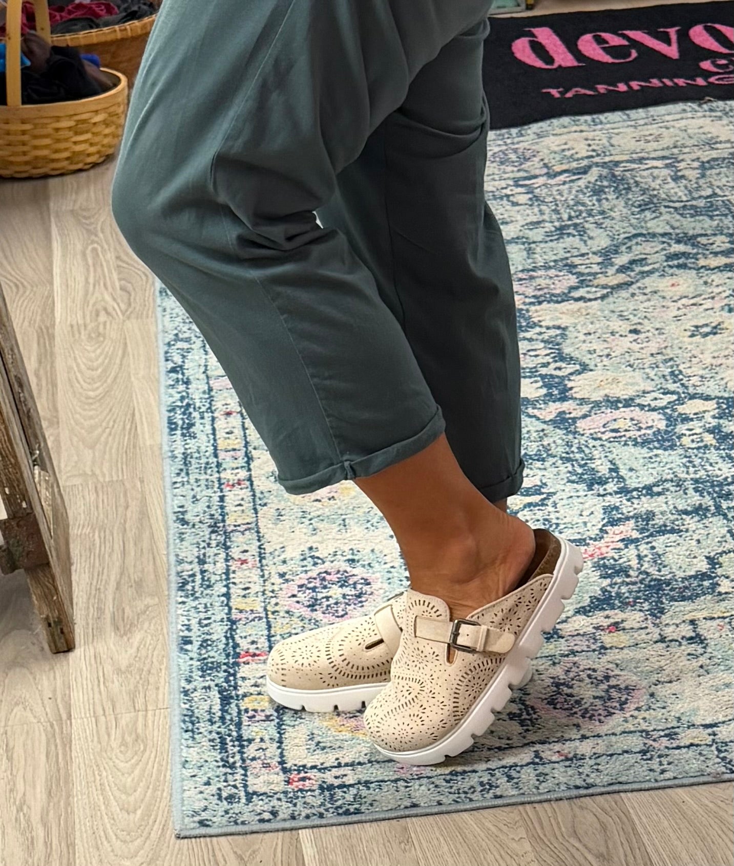 A person wearing beige perforated slip-on shoes with a buckle strap and dark green cropped pants, standing on a distressed blue and white patterned rug in a store.