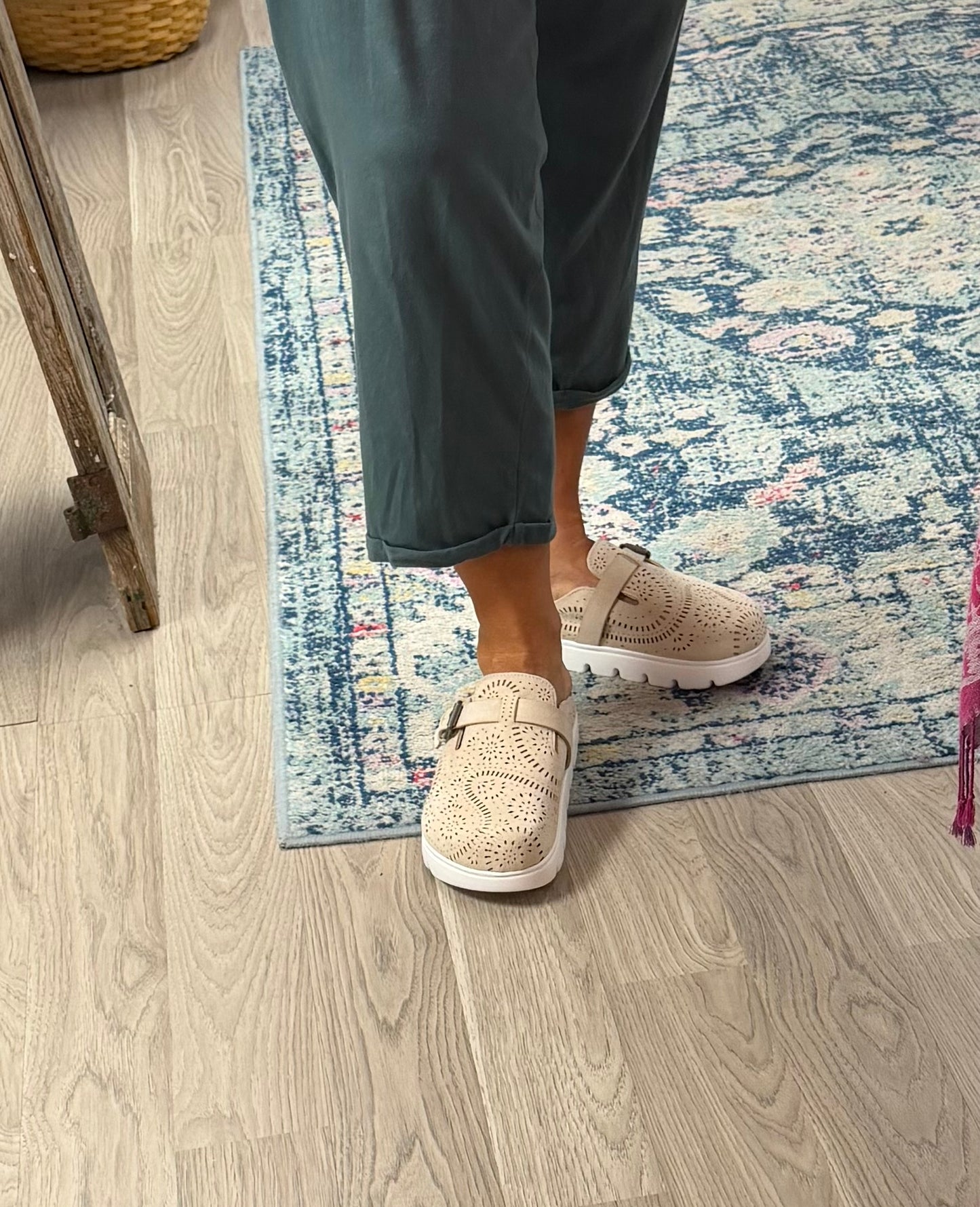 A person wearing beige perforated slip-on shoes with white soles, standing on a blue and white patterned rug over a light wood floor.