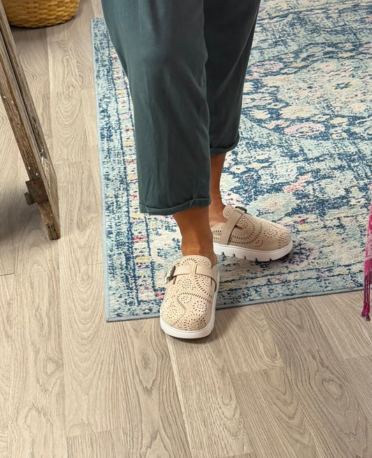 A person wearing beige perforated slip-on shoes with white soles, standing on a blue and white patterned rug over a light wood floor.