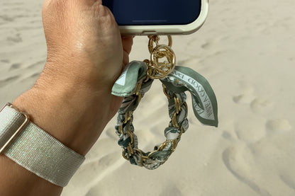 A close-up of a hand holding a phone with a decorative gold-tone chain strap attached. The strap features a mint green and white patterned silk scarf with gold lettering.